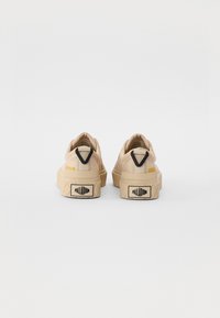 Beige canvas sneakers with a textured surface, featuring a black and yellow accent on the heel and a patterned rubber sole.