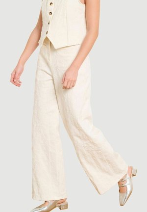 Kobieta ubrana w kremowe szerokie spodnie o fakturze oraz dopasowaną kamizelkę zapinaną na guziki, w połączeniu z metalicznie srebrnymi niskimi butami typu Mary Jane.