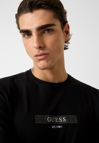 Zwart katoenen T-shirt met een rechthoekige stoffen patch met de tekst "GUESS" en "EST.1981" in witte letters, ronde halslijn en zachte textuur.