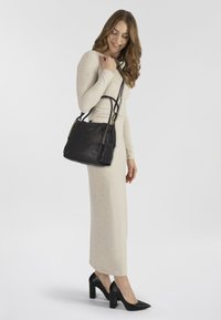 Borsa a tracolla in pelle nera con finitura texture e hardware decorativo. Il modello indossa un lungo abito beige e tacchi neri.