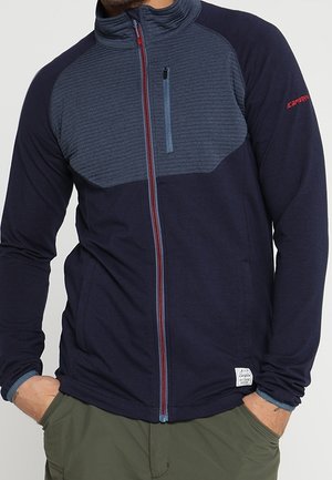 Mężczyzna ubrany w granatowo-szarą polarową kurtkę na suwak z pionową kieszenią na piersi oraz zielone spodnie, ręce w kieszeniach.