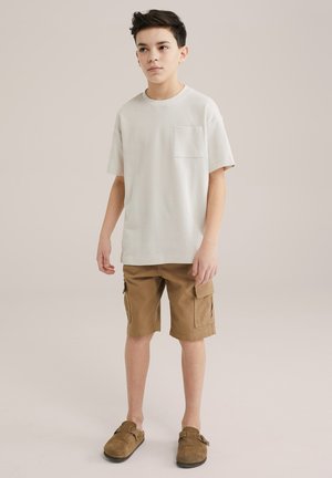 Světle šedé tričko s krátkým rukávem a kapsou, hnědé cargo šortky s bočními kapsami a hnědé slip-on boty. Hladká textura a uvolněný střih.