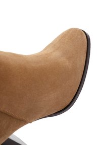 Stivale alla caviglia in suede color cammello con punta affusolata e finitura liscia. La suola è nera con una leggera curvatura nel tallone, mostrando una minima cucitura.