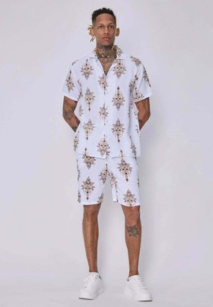 Homme avec des tatouages portant une chemise blanche à manches courtes à motifs et un short assorti, debout, les mains derrière le dos, sur un fond uni.