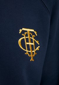 Mörkblå sweatshirt med en guldbroderad logotyp som har sammanflätade bokstäver och dekorativa inslag. Slät textur, design med rund hals.