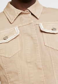 Giacca di jeans beige con due tasche sul petto, bottoni in argento e cuciture dettagliate lungo i bordi delle tasche. Design con colletto e tessuto testurizzato.