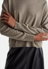 Osoba ubrana w beżowy sweter dzianinowy z prążkowanymi mankietami i dołem, skrzyżowane ramiona na czarnej skórzanej spódnicy.