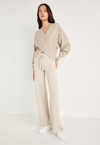 Μπέζ πλεκτή ζακέτα με V λαιμόκοψη και υφασμάτινο σχέδιο, συνδυασμένη με αντίστοιχες φαρδιές παντελόνες και λευκά sneakers. Απαλή και λεία ύφασμα.