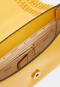 Borsa gialla con una texture liscia, interiori foderati con una tasca con zip e una chiusura a bottone dorato. Decorata con un design intrecciato sulla parte superiore.