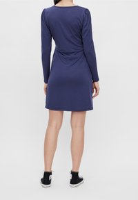 Kvinna som står vänd bort, klädd i en långärmad marinblå klänning och svarta hög-topps sneakers mot en enfärgad vit bakgrund.