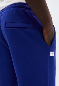Δεξιός γοφός ατόμου που φορά μπλε φόρμα Nike με το χέρι στην μπροστινή τσέπη και ορατή πίσω τσέπη με πατ.