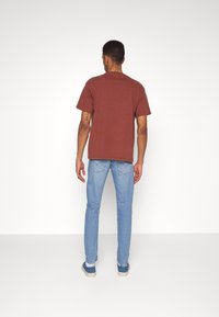 Man som vänder bort med en brun T-shirt, ljusblå jeans och färgglada sneakers mot en enkel vit bakgrund.