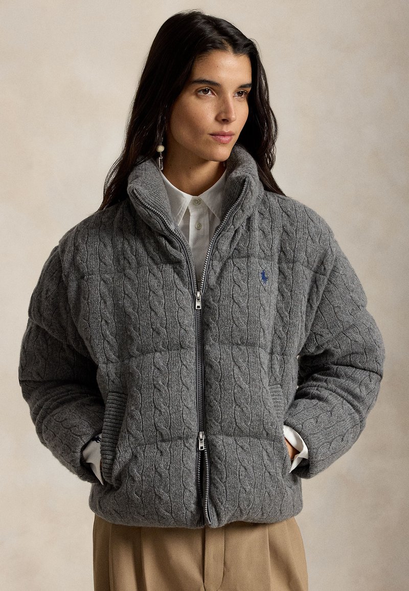 Γυναίκα που φορά γκρι πλεκτό μπουφάν με επένδυση, ψηλό κολάρο, άσπρο πουκάμισο με κουμπιά και μπεζ παντελόνι, στέκεται με τα χέρια στις τσέπες.