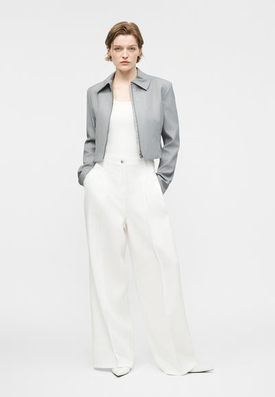 Άτομο στέκεται με τα χέρια στις τσέπες, φορώντας μια γκρι cropped ζακέτα, λευκό τοπ, λευκά παντελόνια φαρδιάς γραμμής και λευκά μυτερά παπούτσια σε απλό φόντο.