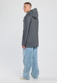 Giacca impermeabile grigia con cappuccio, caratterizzata da un design leggermente svasato e maniche lunghe. Indossata sopra un paio di jeans larghi blu chiaro e sneaker bianche.