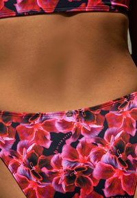 Virágmintás bikini alsó, fekete alappal, nagy piros és rózsaszín virágmintákkal, sima textúrával és fényes logó részlettel.