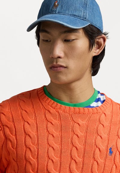 Orange cable-knit sweater with a round neckline, layered over a green striped shirt. A denim cap with a logo sits atop.