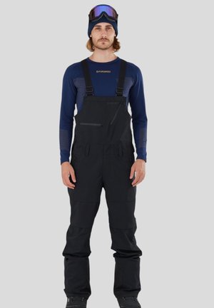 Pantalons de ski noirs en salopette avec des bretelles réglables, des poches zippées et une coupe décontractée, associés à un sous-vêtement à manches longues bleu et des lunettes de ski.