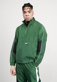 Chaqueta cortavientos verde con cierre frontal, acentos negros y ribete blanco. Presenta el logo "Nike Air" en el pecho. Diseño ligero y deportivo.