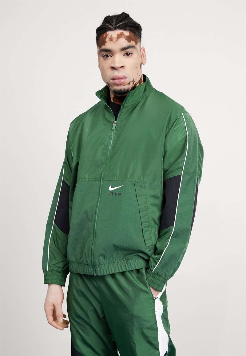 Chaqueta cortavientos verde con cierre frontal, acentos negros y ribete blanco. Presenta el logo "Nike Air" en el pecho. Diseño ligero y deportivo.
