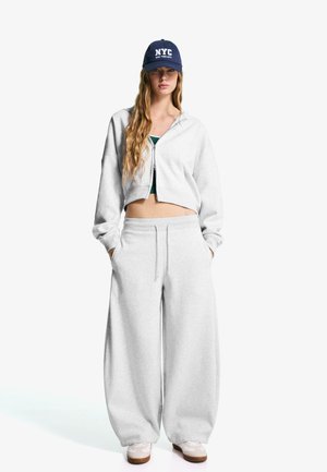 Hellgraue kurze Kapuzenjacke mit Reißverschluss, kombiniert mit lockeren hellgrauen Sweatpants. Weiße Sneakers und eine marineblaue Mütze mit dem Schriftzug "NYC".