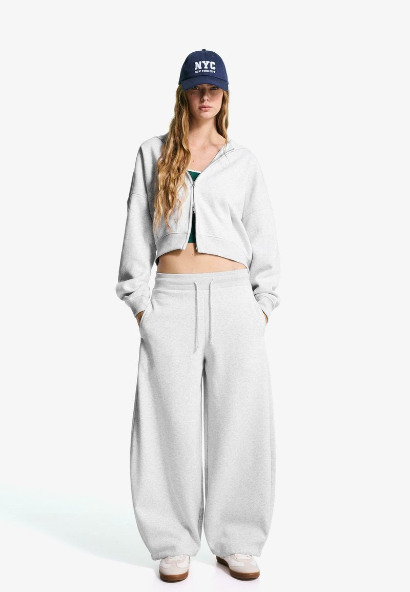 Hanorac scurt de culoare gri deschis, cu fermoar, asociat cu pantaloni de trening largi gri deschis. Adidași albi și o șapcă bleumarin cu textul "NYC".