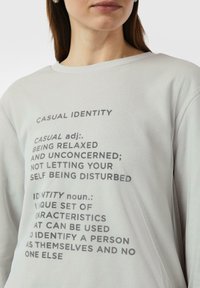 Jasnoszara koszula z długim rękawem z nadrukiem definicji związanych z "casual identity" w ciemnoszarym kolorze. Miękki materiał, luźny krój.