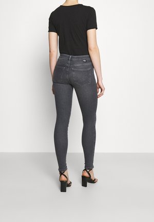 Femme portant un haut noir à manches courtes, un jean skinny gris foncé taille haute, et des sandales noires à talons blocs avec brides, de dos.