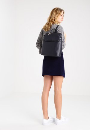 Donna che indossa un maglione grigio, gonna blu navy, sneaker bianche, con uno zaino in pelle nero, in piedi davanti a uno sfondo bianco semplice.