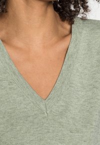 Pull à col en V en tissu tricoté doux de couleur vert clair. Présente un col côtelé et une texture lisse avec des variations subtiles.