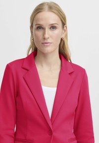 Fuchsia blazer met een getailleerde pasvorm, gesneden revers, sluiting met knopen aan de voorkant en twee zijzakken, gedragen over een witte top.