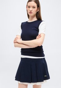 Femme aux cheveux bruns et raides porte un pull sans manches bleu marine par-dessus une chemise blanche à manches courtes et une jupe plissée bleu marine avec un logo vert.