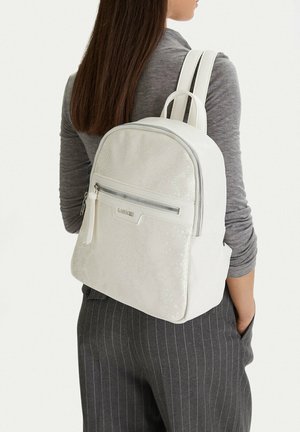 Donna che indossa uno zaino bianco con texture e tasca frontale con cerniera, maglia grigia a maniche lunghe e pantaloni scuri a righe sottili, di spalle con la mano in tasca.