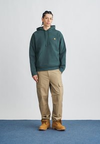 Sudadera con capucha verde oscuro con un gran bolsillo delantero, pantalones cargo beige y botas de trabajo marrones, de pie sobre una superficie alfombrada en azul.