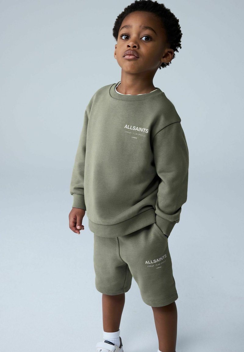 Oliivinvihreä collegepaita- ja shortsiasu, pehmeää kangasta, rento istuvuus, jossa valkoinen logoteksti. Yhdistetty valkoisiin lenkkareihin.