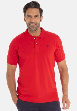 Homme souriant portant un polo à manches courtes rouge avec un petit logo sombre sur la poitrine gauche et un pantalon foncé, les mains en partie dans les poches.