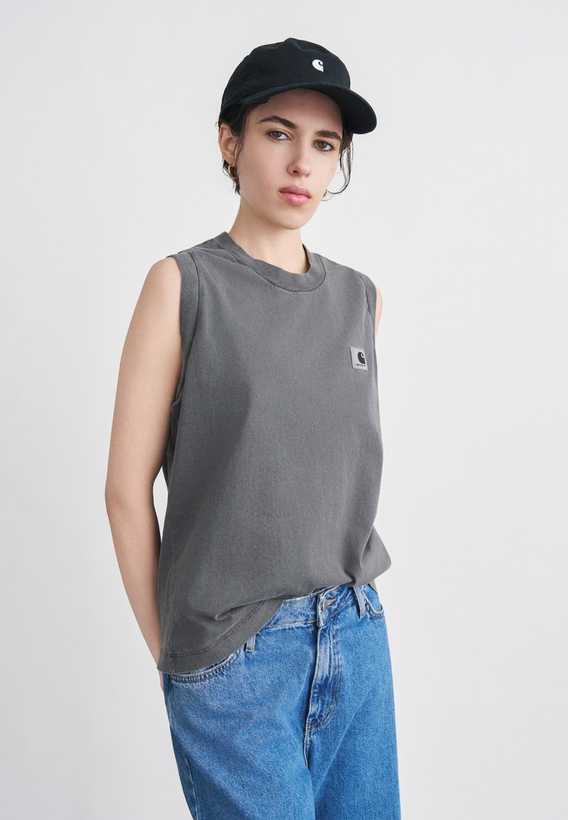 Young person wearing a black cap, sleeveless gray shirt with logo, and blue jeans, standing against a plain light background.