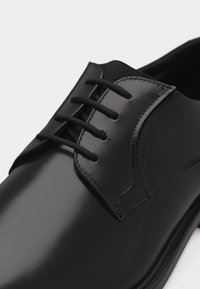 Μαύρο γυαλιστερό δερμάτινο παπούτσι επίσημου τύπου με κλειστό κορδόνι και τρεις οπές για κορδόνια σε λευκό φόντο.