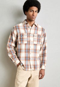Plaid flannel shirt in cream, blue, and rust colors; features button-down design, two front pockets, and long sleeves.