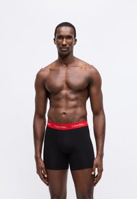 Fekete boxer alsónadrág piros "Calvin Klein" logós derékszíjjal, sima anyagból készült, testhezálló fazonnal és combközép hosszúsággal.