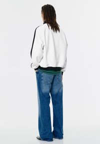 Person med lange dreadlocks iført hvid jakke med sort kant, grøn skjorte, løse blå jeans og sorte sko, står og vender ryggen til.