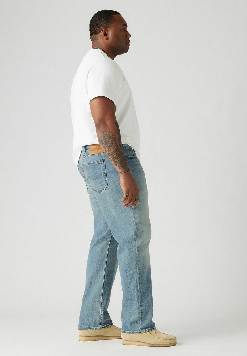 Homme avec un bras tatoué portant un t-shirt blanc, un jean bleu clair, des chaussures beiges et une bague, se tenant de profil contre un fond uni.