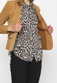 Henkilö säätämässä avointa ruskeaa bleiseriä leopardi-kuosisen nappipaidan ja mustien housujen päällä tasaisessa taustassa.