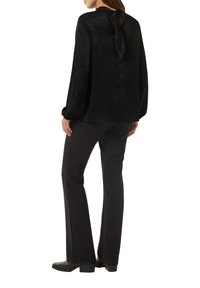 Schwarze Bluse mit langen Ärmeln und einer Schleife am Rücken; glatte, glänzende Textur. Kombiniert mit schmal geschnittenen schwarzen Hosen und Knöchelstiefeln.
