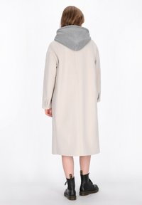 Lungo cappotto beige con cappuccio grigio. Texture liscia, vestibilità rilassata e maniche con bottoni. Indossato sopra stivali neri con lacci.