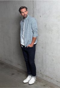 Kostkovaná modro-bílá košile, pod ní bílý tričko, tmavé džíny a bílé tenisky s černými detaily. Betónové pozadí.