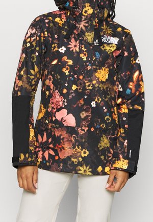 Veste à capuche noire avec imprimé floral multicolore, coudières noires, logo sur la poitrine et bracelets ajustables aux poignets, portée avec un pantalon blanc.