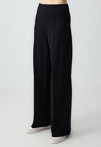 Pantalons larges noirs en tissu lisse avec taille haute, dotés de coutures devant et associés à des baskets blanches.