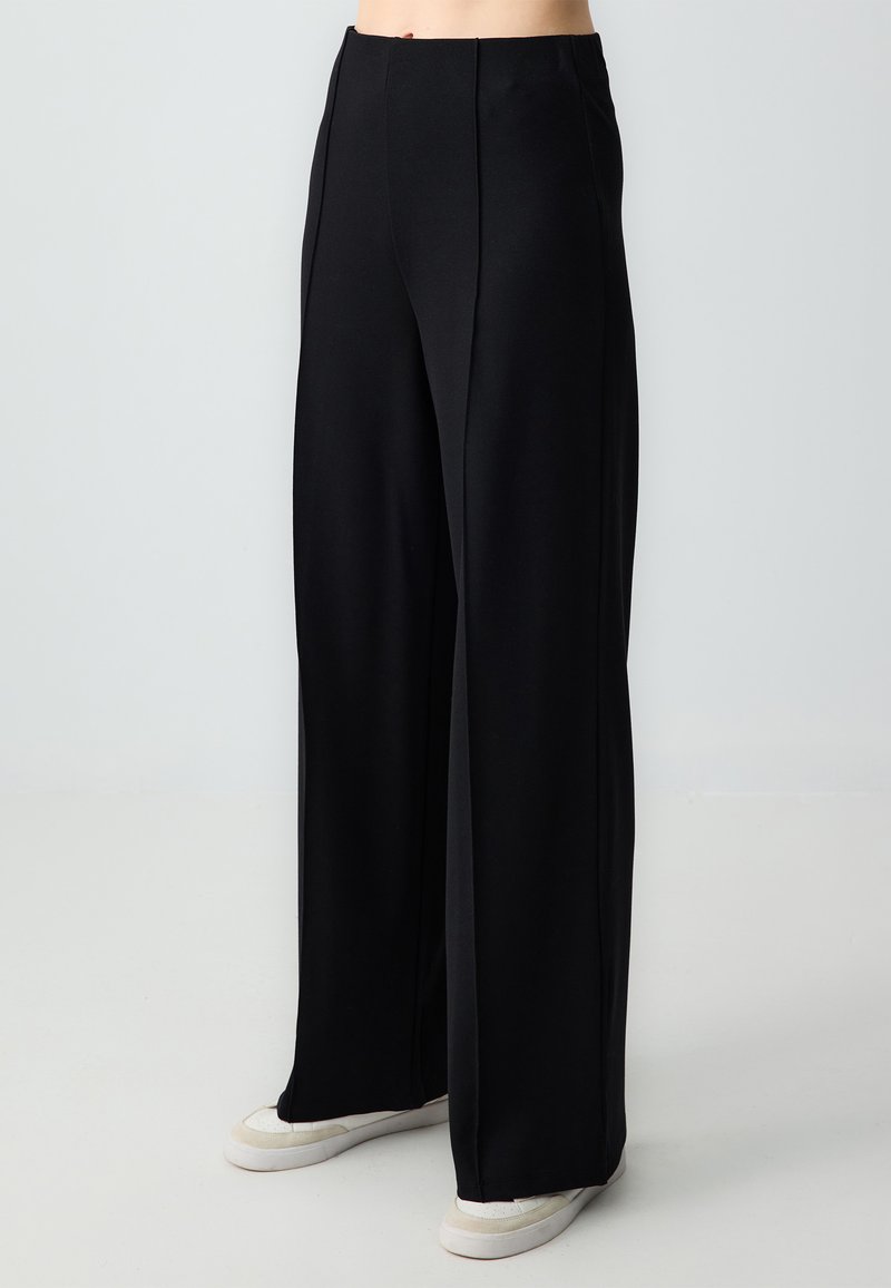 Pantalons larges noirs en tissu lisse avec taille haute, dotés de coutures devant et associés à des baskets blanches.