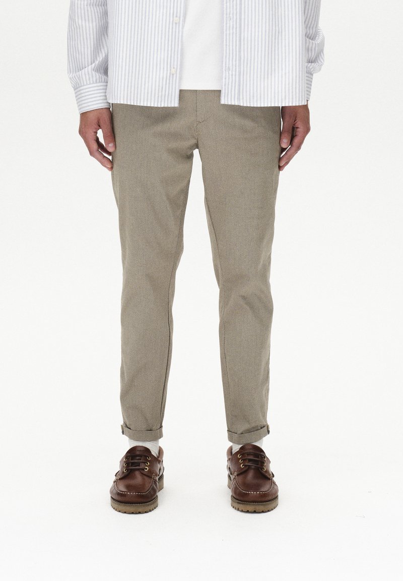 Pantalones de algodón beige con corte ajustado, con los puños enrollados, combinados con zapatos de cuero marrón y una camisa de botones a rayas.
